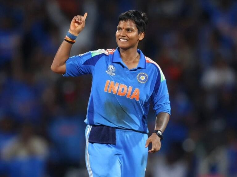 क्रिकेट मैदान में नीली भारतीय टीम की जर्सी पहने दीप्ति शर्मा, जो मुस्कुराते हुए एक हाथ की उंगली ऊपर की ओर उठाए हुए हैं। पीछे स्टेडियम और दर्शकों की धुंधली पृष्ठभूमि दिखाई देती है।