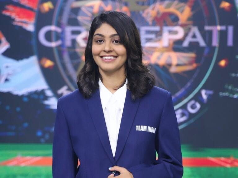 KBC स्टूडियो सेट में खड़ी एक महिला क्रिकेटर हरलीन देओल नीला रंग का ब्लेज़र और सफेद शर्ट पहनी है। ब्लेज़र पर “TEAM INDIA” लिखा है। वे मुस्कुराते हुए सामने देख रही हैं
