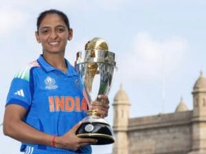 नीली भारतीय क्रिकेट टीम की जर्सी पहने महिला खिलाड़ी, हरामप्रीत कौर मुस्कुराते हुए अपने हाथों में विश्व कप की ट्रॉफी पकड़े हुए हैं। पृष्ठभूमि में खुला आसमान और ऐतिहासिक इमारत दिखाई देती है।
