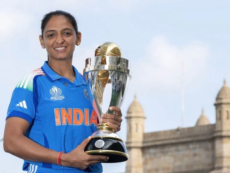 नीली भारतीय क्रिकेट टीम की जर्सी पहने महिला खिलाड़ी, हरामप्रीत कौर मुस्कुराते हुए अपने हाथों में विश्व कप की ट्रॉफी पकड़े हुए हैं। पृष्ठभूमि में खुला आसमान और ऐतिहासिक इमारत दिखाई देती है।