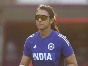 क्रिकेट मैदान में नीली भारतीय टीम की ट्रेनिंग जर्सी पहने स्मृति मंधाना, जिन्होंने स्पोर्ट्स सनग्लासेस लगाए हुए हैं। उनके बाल पीछे बंधे हैं और वे एकाग्र भाव के साथ सामने की ओर देख रही हैं, जबकि पृष्ठभूमि धुंधली दिखाई देती है।