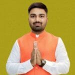 Manish Kashyap, जिसने सफेद कुर्ते के ऊपर नारंगी रंग की नेहरू जैकेट पहनी है। वह हाथ जोड़कर नमस्ते की मुद्रा में कैमरे की ओर देख रहे है और कलाई में घड़ी बंधी है।