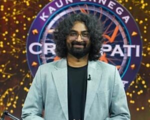 ग्रे सूट और काले टी-शर्ट में रवि गुप्ता KBC शो के मंच पर मुस्कुराते हुए खड़े हैं