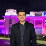 रात के समय रोशनी से सजी एक भव्य इमारत के सामने खड़े शुभांकर मिश्रा, जिन्होंने काले रंग का ब्लेज़र और गहरे रंग की शर्ट पहनी है। वह मुस्कुराते हुए कैमरे की ओर देख रहे है और पीछे बैंगनी व गुलाबी रोशनी से जगमगाती इमारत दिखाई दे रही है।