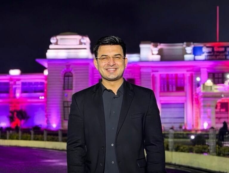 रात के समय रोशनी से सजी एक भव्य इमारत के सामने खड़े शुभांकर मिश्रा, जिन्होंने काले रंग का ब्लेज़र और गहरे रंग की शर्ट पहनी है। वह मुस्कुराते हुए कैमरे की ओर देख रहे है और पीछे बैंगनी व गुलाबी रोशनी से जगमगाती इमारत दिखाई दे रही है।