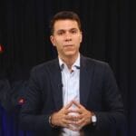 ब्लेज़र और शर्ट पहने tarun Vig (तरुण विग) काले पर्दे के सामने खड़ा होकर बोलते हुए संकेत करता हुआ।