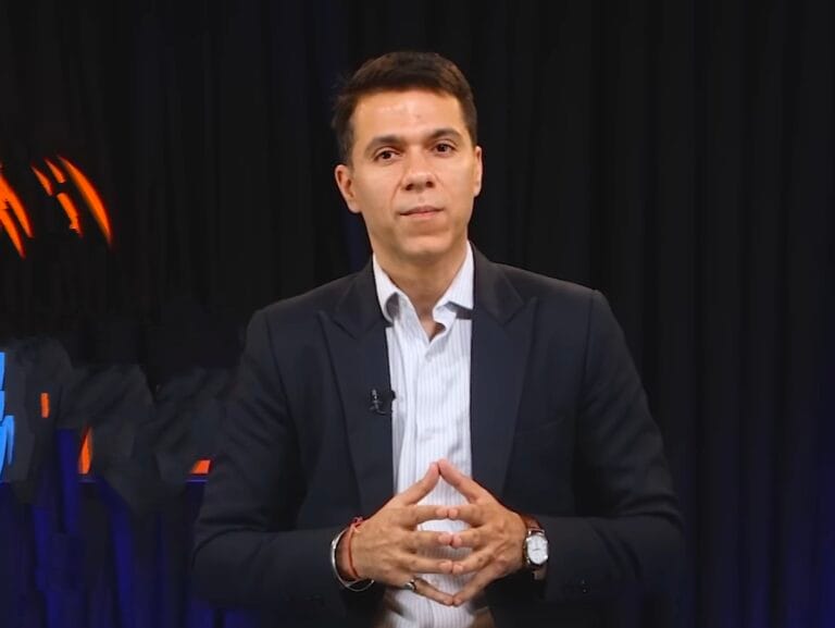 ब्लेज़र और शर्ट पहने tarun Vig (तरुण विग) काले पर्दे के सामने खड़ा होकर बोलते हुए संकेत करता हुआ।